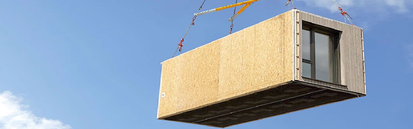 Das Bild zeigt ein vorgefertigtes Modul oder Container, das von einem Kran in die Luft gehoben wird. Dieses Modul scheint Teil eines modularen Bauprojekts zu sein, bei dem vorgefertigte Bauabschnitte verwendet werden, um einen kosteneffizienten und schnellen Aufbau von Gebäuden zu ermöglichen. Das Modul ist aus Holz gefertigt und hat wenigstens eines Fenster. Solche Module werden häufig in der Bauweise von Wohn- und Bürogebäuden oder bei temporären Einrichtungen verwendet.