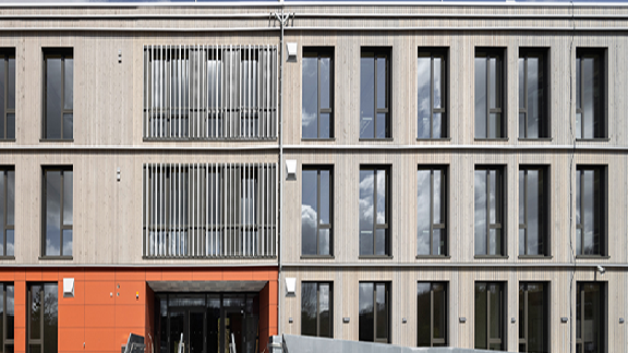 Das Bild zeigt ein modernes Gebäude mit einer Fassade aus Beton und Glas. Es ist ein vierstöckiges Bauwerk mit mehreren großen, rechteckigen Fenstern, die scheinbar symmetrisch angeordnet sind. Die unteren zwei Stockwerke haben zusätzlich eine vertikale Struktur aus Rippen oder Lamellen zwischen den Fenstern, möglicherweise für Sonnenschutz oder ästhetische Zwecke. Der Eingang scheint durch eine belebtere Farbkombination hervorgehoben zu sein, möglicherweise in rot oder orange, was einen Kontrast zur kühlen, grauen Betonfassade bietet.
Dieses Design deutet auf ein Bauwerk hin, das sowohl funktional als auch ästhetisch ansprechend gestaltet ist, möglicherweise eine Institution wie eine Schule, ein Bürogebäude oder ein Verwaltungsgebäude. Die Architektur spiegelt einen modernen und minimalistischen Stil wider, der in urbanen Gebieten häufig anzutreffen ist.