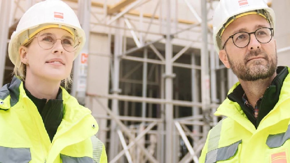 Das Bild zeigt zwei Personen in Sicherheitskleidung und Bauhelmen vor einem Baugerüst. Sie wirken wie Bauarbeiter oder Ingenieure auf einer Baustelle.