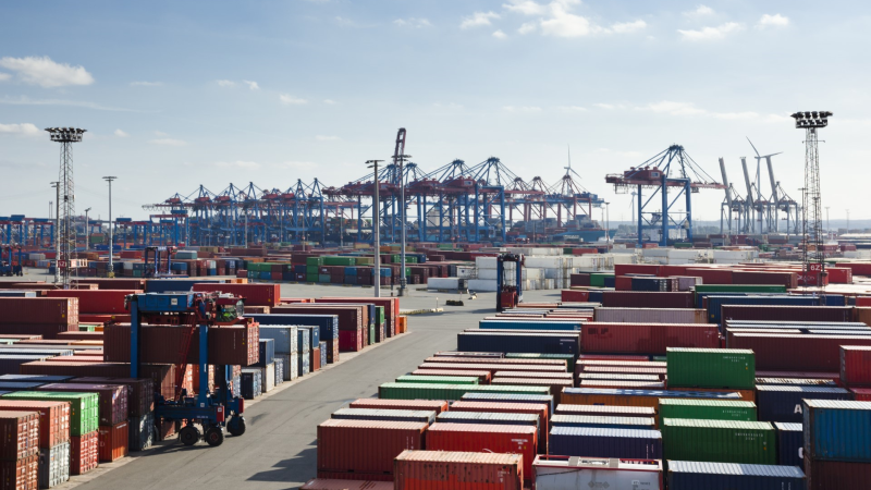 Das Bild zeigt einen Containerhafen mit vielen gestapelten Containern und Kränen im Hintergrund. Es handelt sich um eine typische Hafenanlage, die für den Umschlag von Frachtcontainern verwendet wird. Diese Umgebung ist charakteristisch für einen industriellen und logistischen Bereich, in dem der internationale Warenverkehr abgewickelt wird.