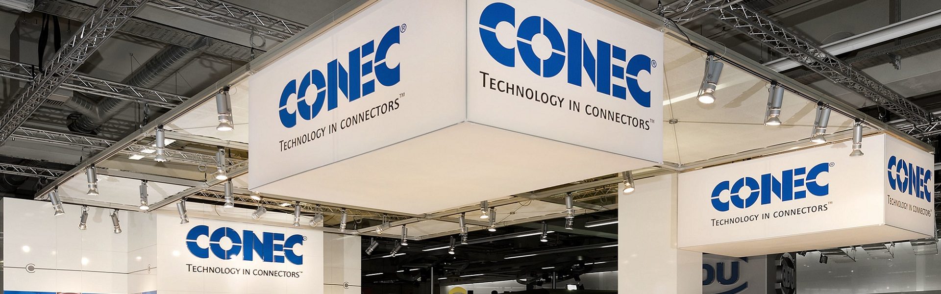 Das Bild zeigt einen Messestand oder eine Ausstellungsfläche eines Unternehmens namens CONEC. CONEC scheint in der Technologiebranche tätig zu sein, wie aus dem Slogan „Technology in Connectors“ ersichtlich ist, der auf den Schildern des Standes zu sehen ist. Die Schriftzüge auf den Bannern und Schildern der Firma CONEC sind gut sichtbar, und die Anordnung des Standes deutet auf eine professionelle Darstellung ihrer Produkte und Dienstleistungen in einer Messeumgebung hin.