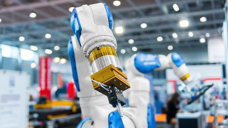 Das Bild zeigt einen industriellen Roboterarm in einer technologischen Umgebung, vermutlich in einer Fabrik oder während einer Messe für industrielle Automation.
Beschreibung des Bildes:
1. **Roboterarme:** Zwei weiße und blaue Roboterarme stehen im Vordergrund des Bildes. Der Fokus liegt auf dem vorderen Arm, der gut sichtbar ist und mit einem speziellen Werkzeug ausgestattet ist.
2. **Werkzeug:** Am Ende des Roboterarms ist ein Werkzeug oder eine Greifvorrichtung befestigt, das möglicherweise für spezielle Bearbeitungs-, Montage- oder Präzisionsarbeiten verwendet wird.
3. **Umgebung:** Der Hintergrund deutet auf eine gut beleuchtete Halle hin, wahrscheinlich eine industrielle Umgebung oder eine Messe, wo verschiedene technologische Geräte und Lösungen präsentiert werden.
4. **Technologische Entwicklung:** Das Bild vermittelt ein Gefühl von fortschrittlicher Technologie und zeigt, wie moderne Maschinen zur Automatisierung eingesetzt werden könnten.
5. **Unschärfe im Hintergrund:** Die leichte Unschärfe im Hintergrund deutet auf Aktivität und weitere Ausstellungsstände oder Maschinen hin, jedoch konzentriert sich das Bild auf den Roboterarm selbst als Hauptblickfang.