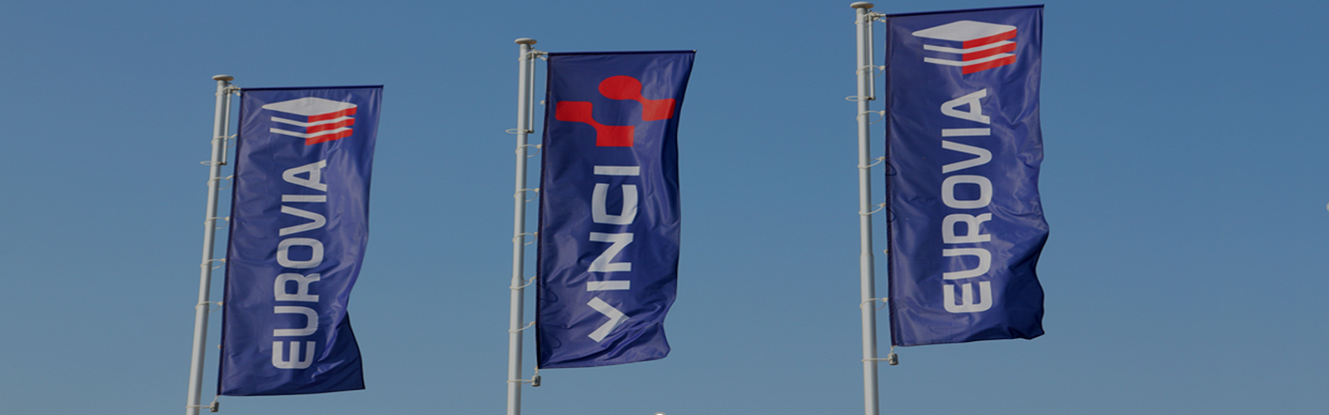 Das Bild zeigt drei Fahnen, die im Wind wehen, jeweils auf einem Mast befestigt. Die Fahnen tragen das Logo und den Namen von EUROVIA und VINCI.

**EUROVIA**:
- Eine Marke von einem Unternehmen, das hauptsächlich im Bereich Straßenbau, Verkehrsinfrastruktur und Dienstleistungen tätig ist.
- Teil des Baukonzerns VINCI.

**VINCI**:
- Ein weltweit führendes Konzern im Bauwesen und für Konzessionen.
- Entwickelt und realisiert Projekte in den Bereichen Bau, Infrastruktur, Energie, Immobilien sowie im Bereich Dienstleistungen.

**Gesamtbetrachtung**:
- Die Fahnen symbolisieren eine Baustelle oder einen Unternehmensstandort von VINCI und EUROVIA, wahrscheinlich in der Nähe von Bau- oder Infrastrukturprojekten.
- Das klare Design und die Erwähnung der Marken verweisen auf die umfassenden Bau- und Ingenieurdienstleistungen, die die Unternehmen anbieten.