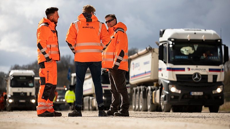 Das Bild zeigt drei Männer in orangefarbenen Sicherheitsjacken, die auf einer Baustelle stehen. Sie scheinen sich zu unterhalten oder zu beraten. Im Hintergrund sind mehrere große Lastwagen zu sehen, darunter ein Mercedes-Benz-Lkw. Die Männer tragen eine Arbeitskleidung, die für Baustellen typisch ist, um ihre Sichtbarkeit und Sicherheit zu erhöhen, speziell durch die leuchtenden Farben der Jacken. Die Szene vermittelt den Eindruck einer organisierten und geschäftigen Baustellenumgebung, möglicherweise im Bereich des Straßenbaus oder der Infrastruktur.