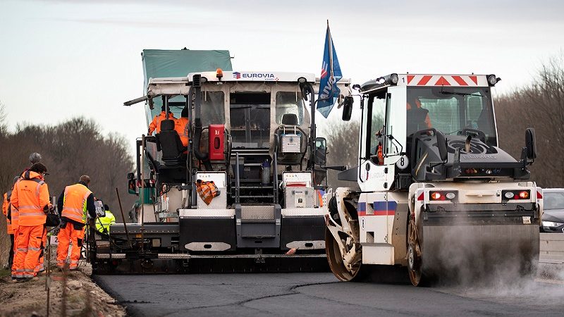 Das Bild zeigt eine Straßenbau-Szene. Im Mittelpunkt stehen mehrere Bauarbeiter, die an einem Straßenbauprojekt arbeiten. Sie sind mit leuchtend orangefarbenen Sicherheitskleidung ausgestattet, was darauf hinweist, dass sie in einem Arbeitsbereich tätig sind. 

Der asphaltierte Bereich wird von spezifischen Straßenbaugeräten bearbeitet:

1. **Asphaltfertiger**: Das große Gerät in der Mitte des Bildes ist ein Asphaltfertiger oder Straßenfertiger, der frischen Asphalt aufträgt und ihn gleichmäßig auf der Straßenoberfläche verteilt.

2. **Straßenwalze**: Rechts von dem Asphaltfertiger ist eine Straßenwalze zu sehen, die die Aufgabe hat, den Asphalt zu verdichten, um eine glatte und stabile Straßenoberfläche zu schaffen.

Hintergrund:
- Die Szene spielt sich in einer ländlichen oder vorstädtischen Umgebung ab, da im Hintergrund Baumreihen zu sehen sind.

Zusammenfassend zeigt das Bild den Prozess des Straßenbaus, bei dem Asphalt aufgetragen und verdichtet wird, um eine neue oder erneuerte Fahrbahn zu schaffen. Es illustriert die Teamarbeit, die bei solchen Infrastrukturprojekten erforderlich ist.