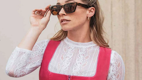 Die abgebildete Person trägt eine rot-weiße Tracht oder traditionelles Dirndl. Das Oberteil besteht aus einem roten Korsett mit weißer Spitze darunter, typischerweise elegant und oft für Anlässe wie Oktoberfeste oder andere traditionelle Feiern getragen. Die Person hält eine Sonnenbrille in der Hand, was auf Modebewusstsein oder Vorbereitung auf sonniges Wetter hinweisen könnte.
