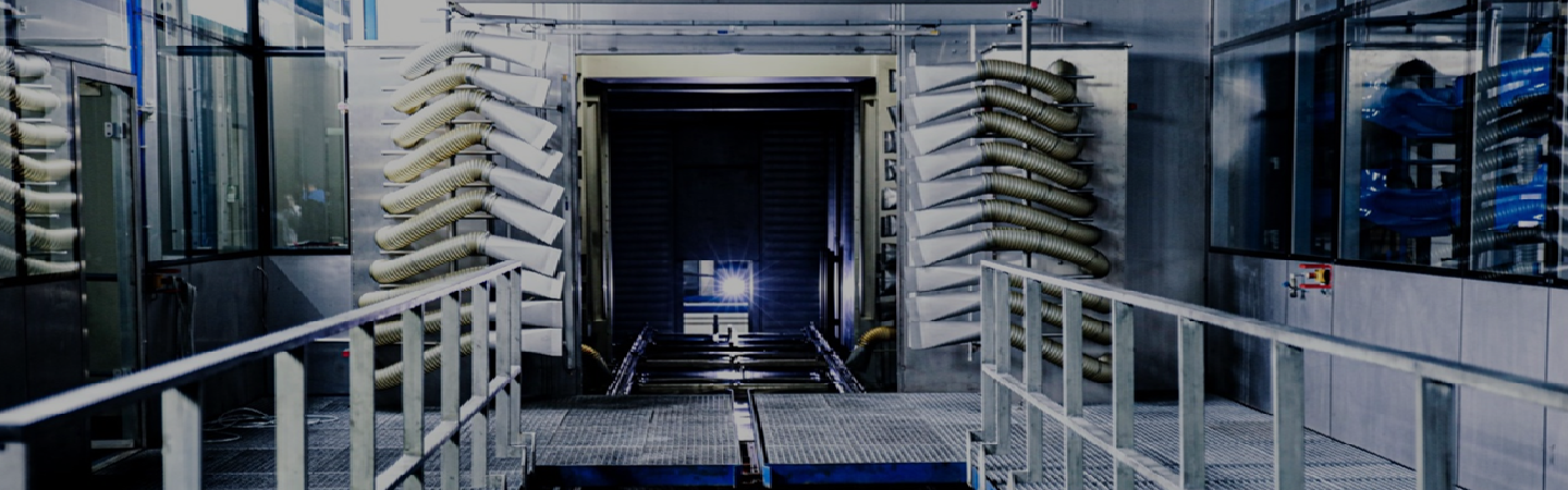 Das Bild zeigt ein industrielles Umfeld, vermutlich eine Produktionsanlage oder Fabrik. Der zentrale Bereich hat eine Konstruktion mit Schienen oder einem Förderband in der Mitte, das scheinbar in einen Tunnel oder eine Kammer führt. Auf beiden Seiten sind große, vertikale Strukturen zu sehen, die möglicherweise Regale oder Halterungen für Produkte sind. Die kühle Farbgebung und das technische Design des Raums lassen darauf schließen, dass dies ein Ort für industrielle Prozesse oder Maschinenarbeit ist. Es könnte sich um eine Anlage zur Produktion, Verarbeitung oder vielleicht sogar Verpackung handeln. Der helle Schein im Hintergrund könnte von Beleuchtung stammen, die für die Arbeitsumgebung typisch ist.**