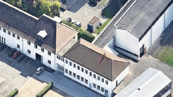 Das Bild zeigt ein Luftbild oder eine Vogelperspektive eines Gewerbegebiets oder Industriekomplexes. Man sieht mehrere Gebäude mit unterschiedlichen Dachformen und Baumaterialien. Das Hauptgebäude scheint ein Büro- oder Verwaltungsgebäude zu sein, da es Fenster und Zugangspunkte auf der Vorderseite hat.
**Merkmale des Bildes:**
1. **Hauptgebäude:**
- Das Hauptgebäude hat ein geneigtes Dach und wirkt wie ein Bürogebäude.
- Es gibt mehrere Fensterreihen, was darauf hinweist, dass dort möglicherweise Büroräume sind.
- Ein Eingang ist auf der Vorderseite zu erkennen.
2. **Nebenstruktur:**
- Neben dem Hauptgebäude steht eine größere Halle oder Lager mit einem Flachdach.
- Diese Struktur könnte als Lager- oder Produktionsstätte genutzt werden.
3. **Umgebung:**
- Der Bereich um die Gebäude ist mit einem gepflasterten Parkplatz ausgestattet.
- Verschiedene Fahrzeuge sind auf dem Gelände geparkt.
- Es gibt möglicherweise eine grüne Zone oder Bepflanzungen in der Umgebung der Gebäude.
4. **Zugang:**
- Das Gelände scheint einen zentralen Zugangspunkt für Fahrzeuge zu haben, der von den umliegenden Straßen erreicht werden kann.
Insgesamt vermittelt das Bild den Eindruck eines funktionellen Gewerbe- oder Industriekomplexes, der für geschäftliche Zwecke genutzt wird. Gebäude-und Geländestruktur weisen auf eine gut organisierte Arbeitsumgebung hin.