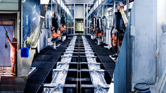 Das Bild zeigt eine industrielle Lackierstraße, die für den Lackierprozess von Fahrzeugen oder Maschinen verwendet wird. Die Lackierstraße ist mit mehreren Robotern ausgestattet, die scheinbar für das automatische Aufbringen von Farbe auf die Oberflächen der zu bearbeitenden Teile zuständig sind. Das gesamte Setup ist darauf ausgelegt, den Lackierprozess effizient und kontrolliert durchzuführen und dabei menschlichen Eingriff zu minimieren.
### Detailbeschreibung:
1. **Automatisierte Roboter:** Mehrere Roboterarme sind entlang der Lackierstraße positioniert, möglicherweise ausgestattet mit Sprühdüsen, um die Farbe präzise und gleichmäßig aufzutragen.
2. **Umgebung:** Die Lackierstraße befindet sich in einem geschlossenen Raum, wahrscheinlich um die Umgebung zu kontrollieren und die Qualität des Lackierprozesses zu gewährleisten.
3. **Sicherheitsausstattung:** Schutzkleidung und möglicherweise eine Absauganlage sind vorhanden, um den Arbeitsschutz zu gewährleisten und die Freisetzung von Dämpfen zu minimieren.
4. **Bodenstruktur:** Der Boden scheint mit Schutzmatten oder Gittern ausgelegt zu sein, um das Ablaufen von überschüssiger Farbe und Lösungsmitteln zu ermöglichen.
Das Bild illustriert die technologische Entwicklung und industrielle Fortschritte bei der Automatisierung von Produktionsprozessen in der Fertigungsindustrie.